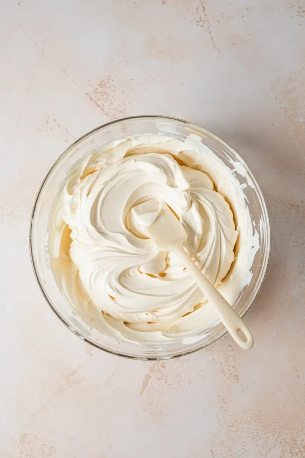 Creamy no-bake cheesecake filling in a mixing bowl with a white spatula, showing smooth swirls and a soft texture on a pastel backdrop.