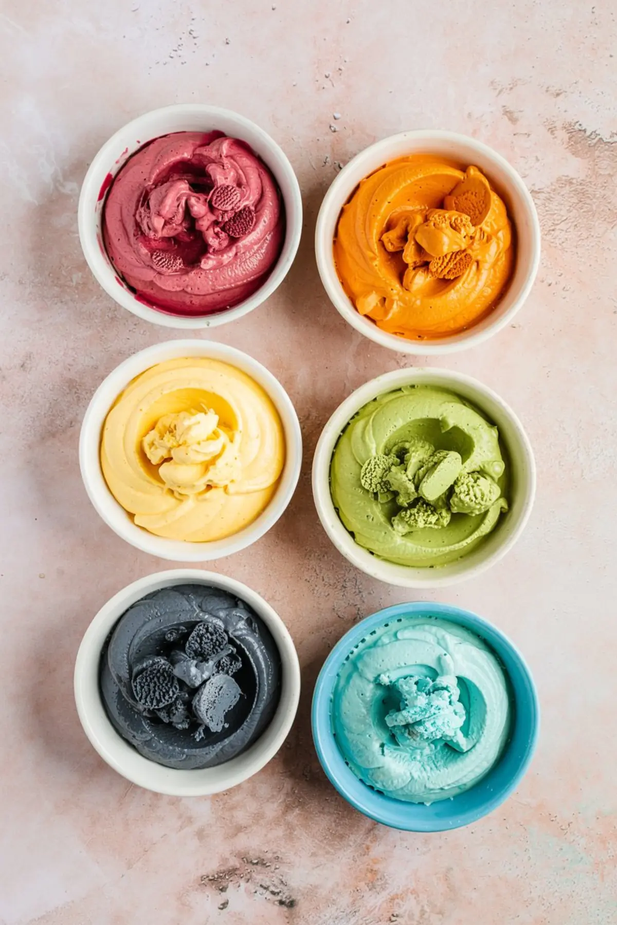 Six small bowls filled with colorful cheesecake layers in red, orange, yellow, green, black, and blue, displayed on a pink surface for a rainbow dessert recipe.
