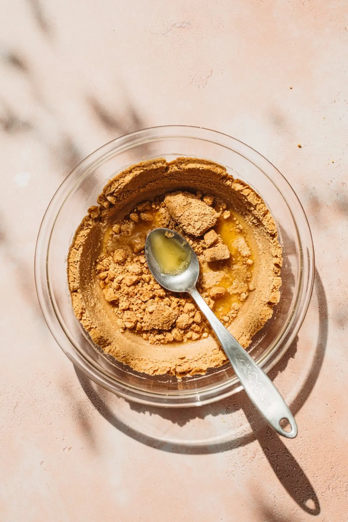 Graham cracker crumbs and melted butter mixture in a clear bowl with a spoon, ready to be mixed for a cheesecake crust, placed on a warm-toned background.