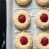 Raspberry coconut thumbprint cookies arranged on a parchment-lined baking tray, topped with whole raspberries and rolled in shredded coconut, with white icing drizzled across the surface.