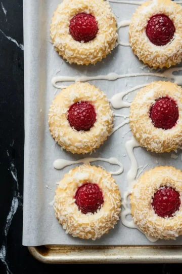 Raspberry coconut thumbprint cookies arranged on a parchment-lined baking tray, topped with whole raspberries and rolled in shredded coconut, with white icing drizzled across the surface.