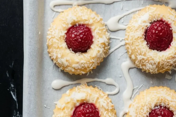 Raspberry coconut thumbprint cookies arranged on a parchment-lined baking tray, topped with whole raspberries and rolled in shredded coconut, with white icing drizzled across the surface.