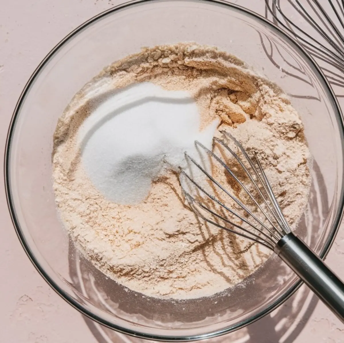 Clear glass bowl filled with a dry mixture of flour and sugar being whisked with a metal balloon whisk, placed on a pale pink background for a baking preparation scene.