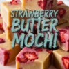 Close-up of sliced strawberry butter mochi squares stacked on a wooden platter, showing the glossy, chewy texture with visible strawberry chunks and bold text overlay describing the dessert.