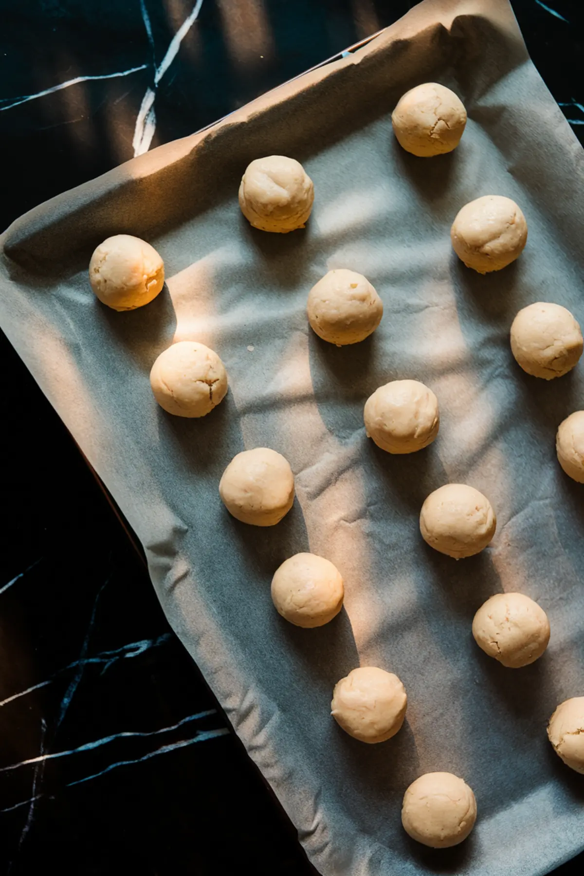 Raw cookie dough balls spaced evenly on a parchment-lined baking tray, ready to be baked, with soft shadows highlighting the texture of the dough on a black background.