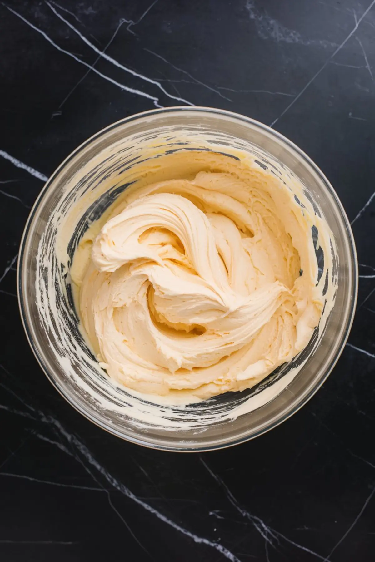 Thick, whipped cookie dough in a glass mixing bowl with a creamy, smooth texture, shown on a black marble countertop.