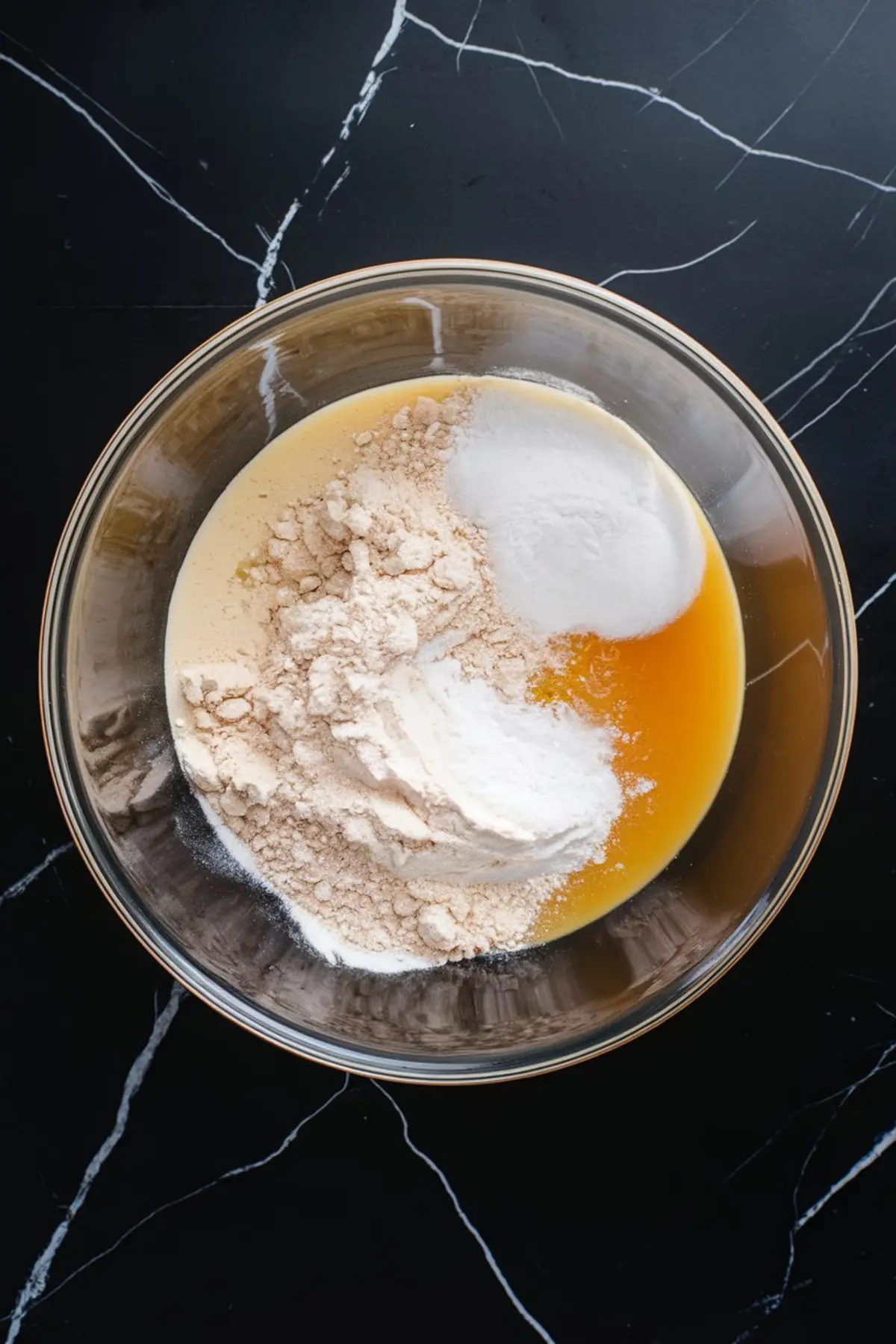 Mixing bowl filled with cookie dough in progress, showing flour, sugar, and wet ingredients like vanilla and oil just before being combined on a black surface.