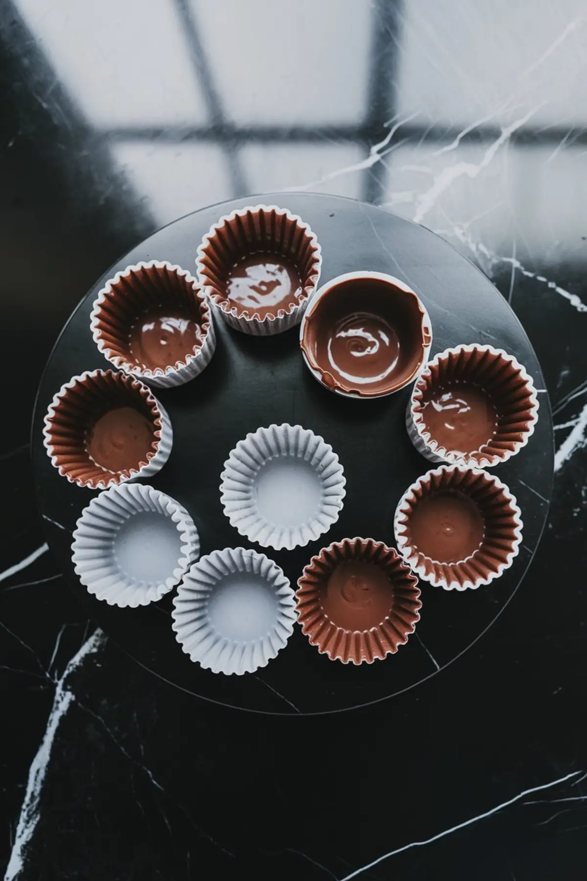 Round black plate holding a circle of paper cupcake liners, some filled with melted chocolate and others empty, on a dark marble surface.