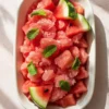 Rectangular white dish filled with watermelon granita, garnished with fresh mint leaves and watermelon wedges, placed on a sunlit textured surface with soft shadows.