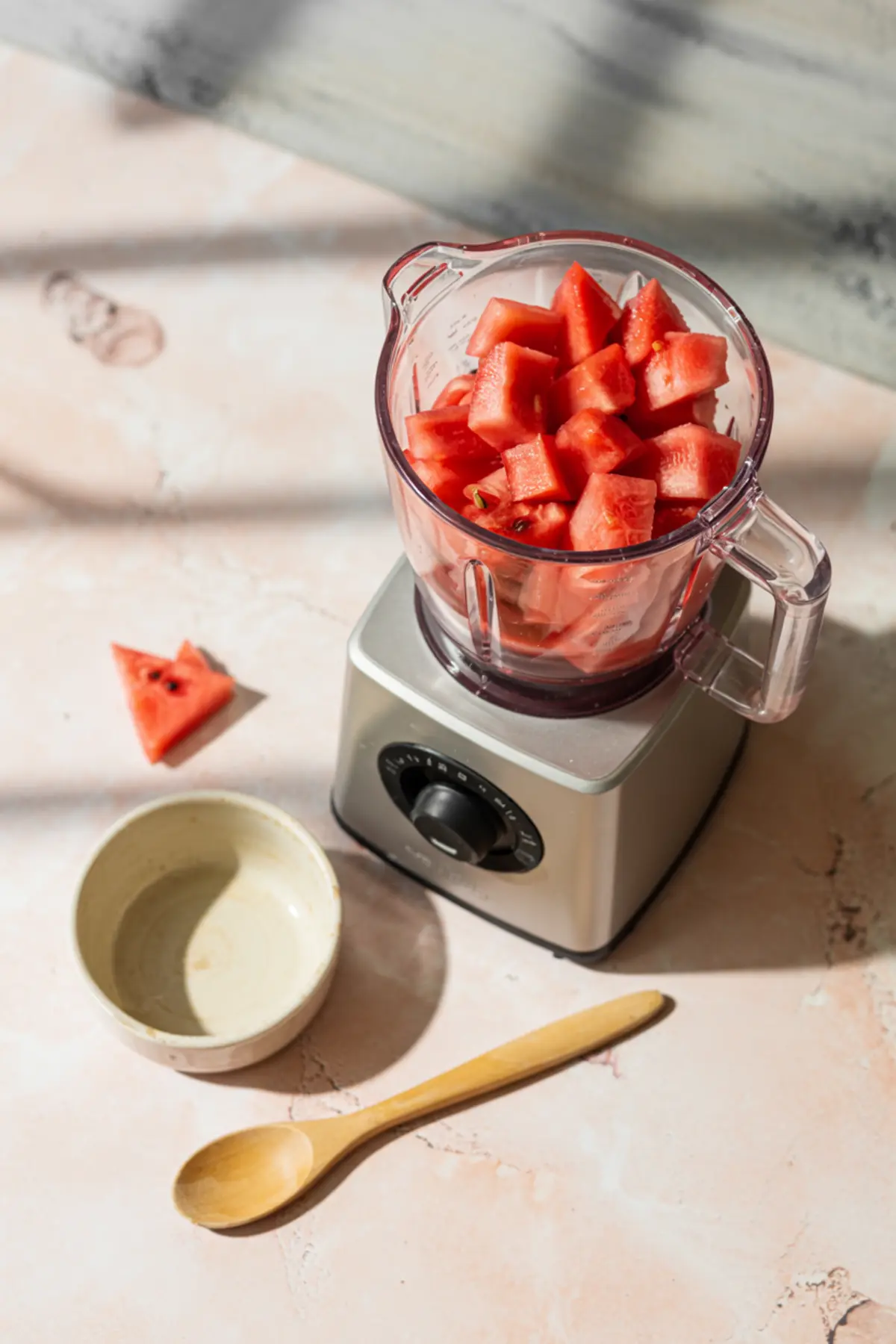 Blender filled with fresh cubed watermelon on a pink marbled surface, with a ceramic bowl, wooden spoon, and a triangle slice of watermelon nearby.