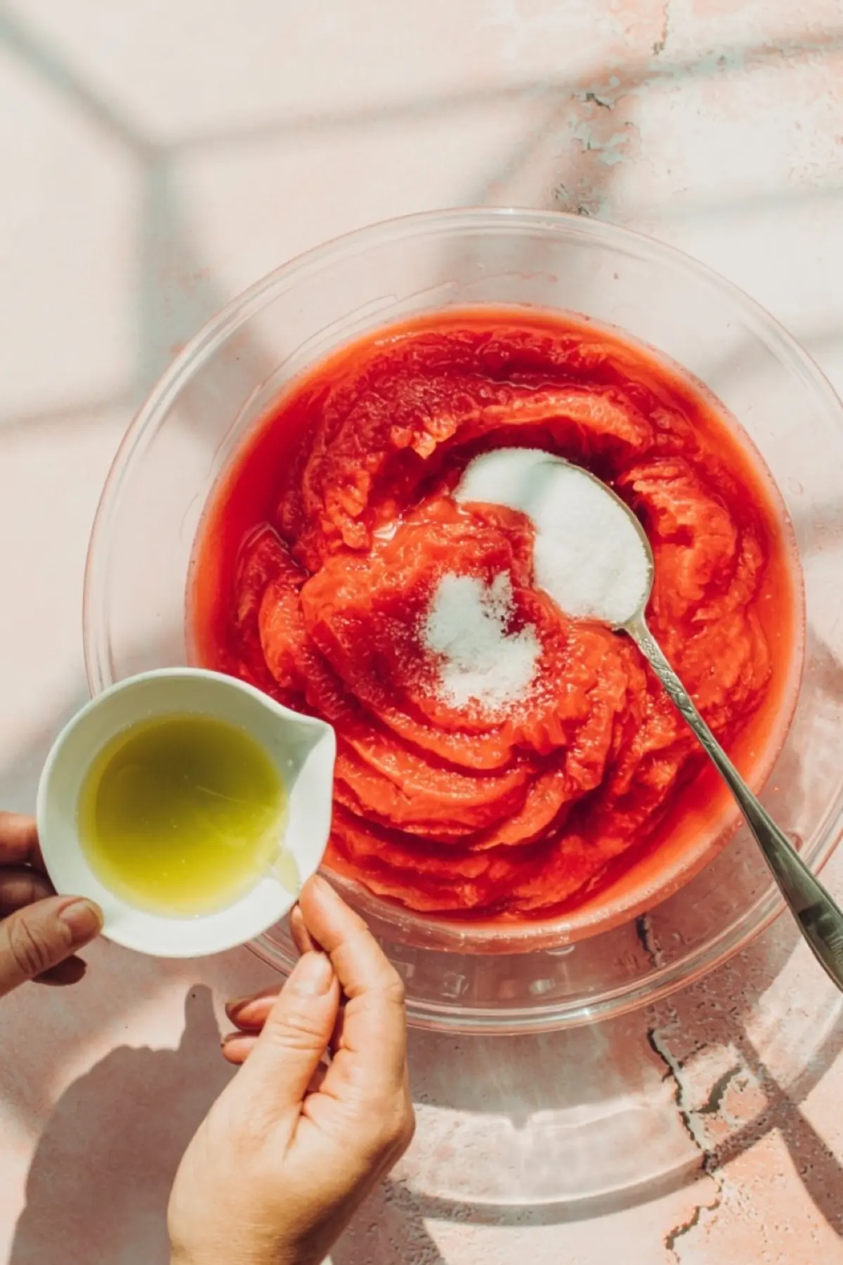 Overhead view of blended watermelon mixture in a clear glass bowl with sugar sprinkled on top, a spoon resting inside, and a hand holding a small pitcher of lemon juice above the bowl.