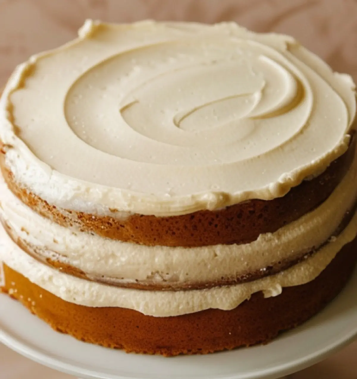 Three-layer white chocolate cake with thick vanilla frosting between layers and a smooth, swirled top. The cake is unfrosted on the sides, highlighting its golden sponge and creamy texture.
