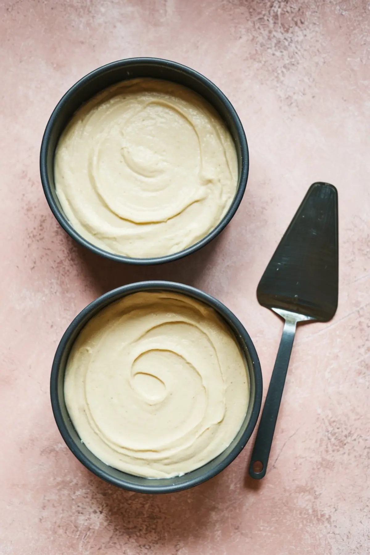 Two round cake pans filled with white chocolate cake batter, each smoothed into a swirl pattern. The pans are placed on a light pink surface beside a metal cake server.
