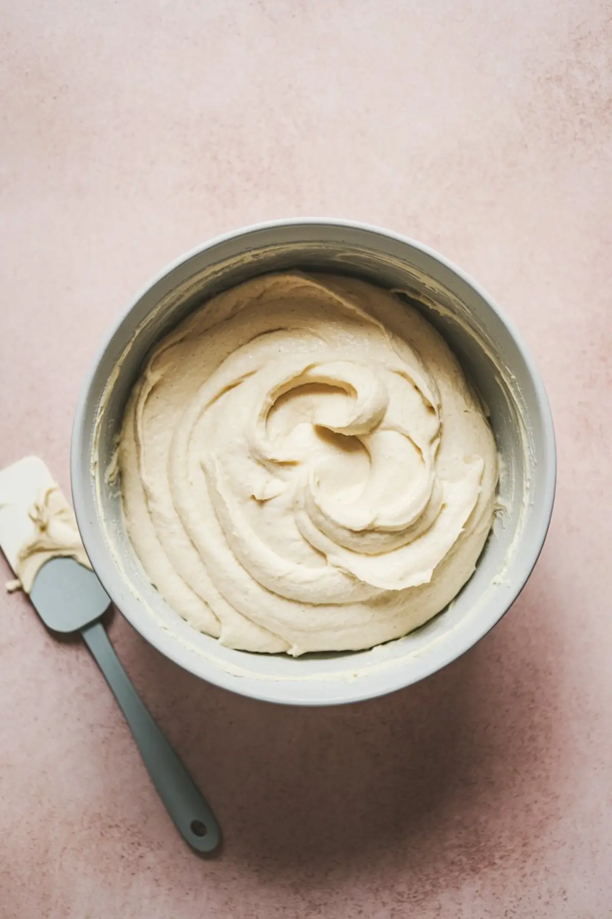 Metal mixing bowl filled with swirled white chocolate cake batter, set on a soft pink background. A spatula with residual batter rests beside the bowl.
