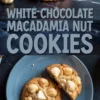 White chocolate macadamia nut cookies on a dark countertop, featuring a stack of golden cookies and a halved cookie on a blue plate. The cookies have a rich, soft texture and are generously filled with whole macadamia nuts and white chocolate pieces.