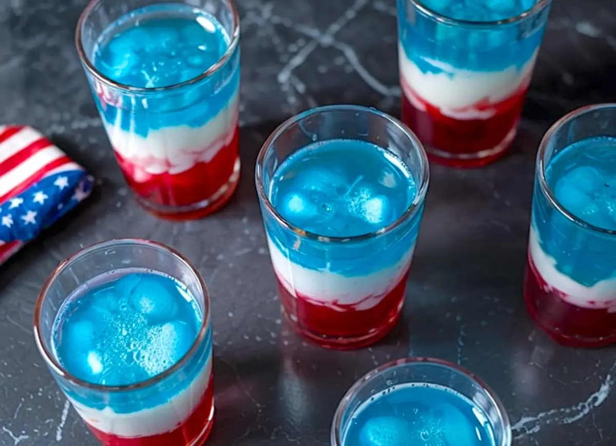 Overhead view of multiple 4th of July layered cocktails with red, white, and blue layers topped with ice cubes, arranged on a black marble surface beside a folded American flag napkin.
