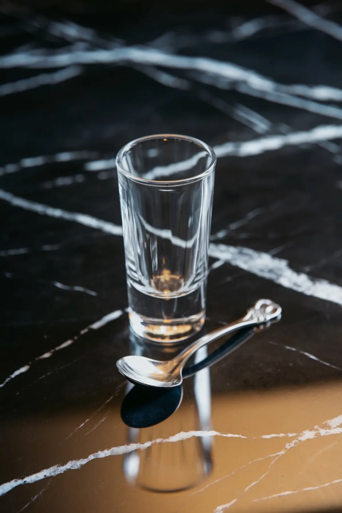 Empty shot glass with a bar spoon on a polished black marble surface, showing preparation setup for mixing layered cocktails.
