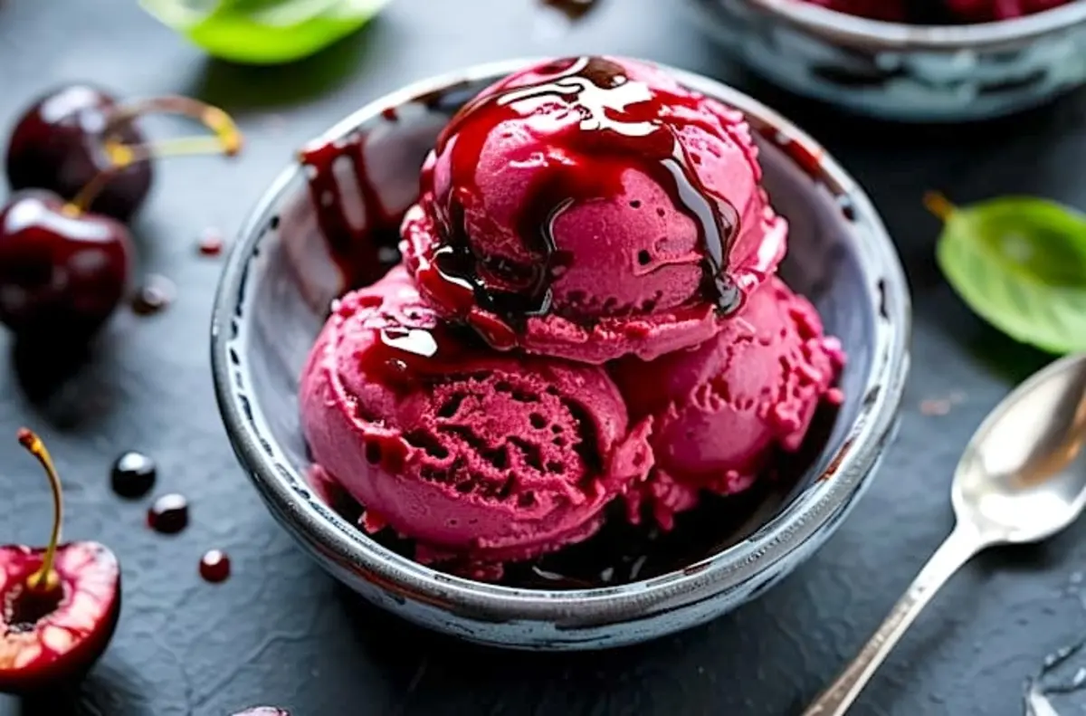 A bowl of black cherry sorbet with scoops drizzled in glossy balsamic glaze, surrounded by fresh cherries and basil leaves on a dark textured background.