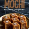 Close-up of golden brown butter mochi cubes topped with flaky sea salt, arranged neatly on a metallic gold plate over a dark marble surface.
