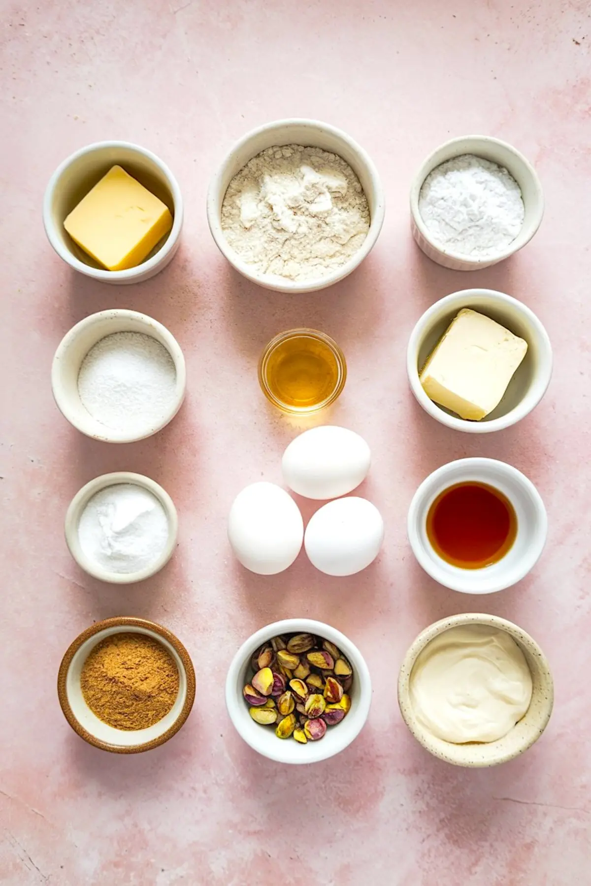 Flat lay of ingredients for brown butter honey pistachio bars, including butter, flour, sugar, eggs, honey, vanilla extract, baking powder, pistachios, cream cheese, and spices on a pink background.