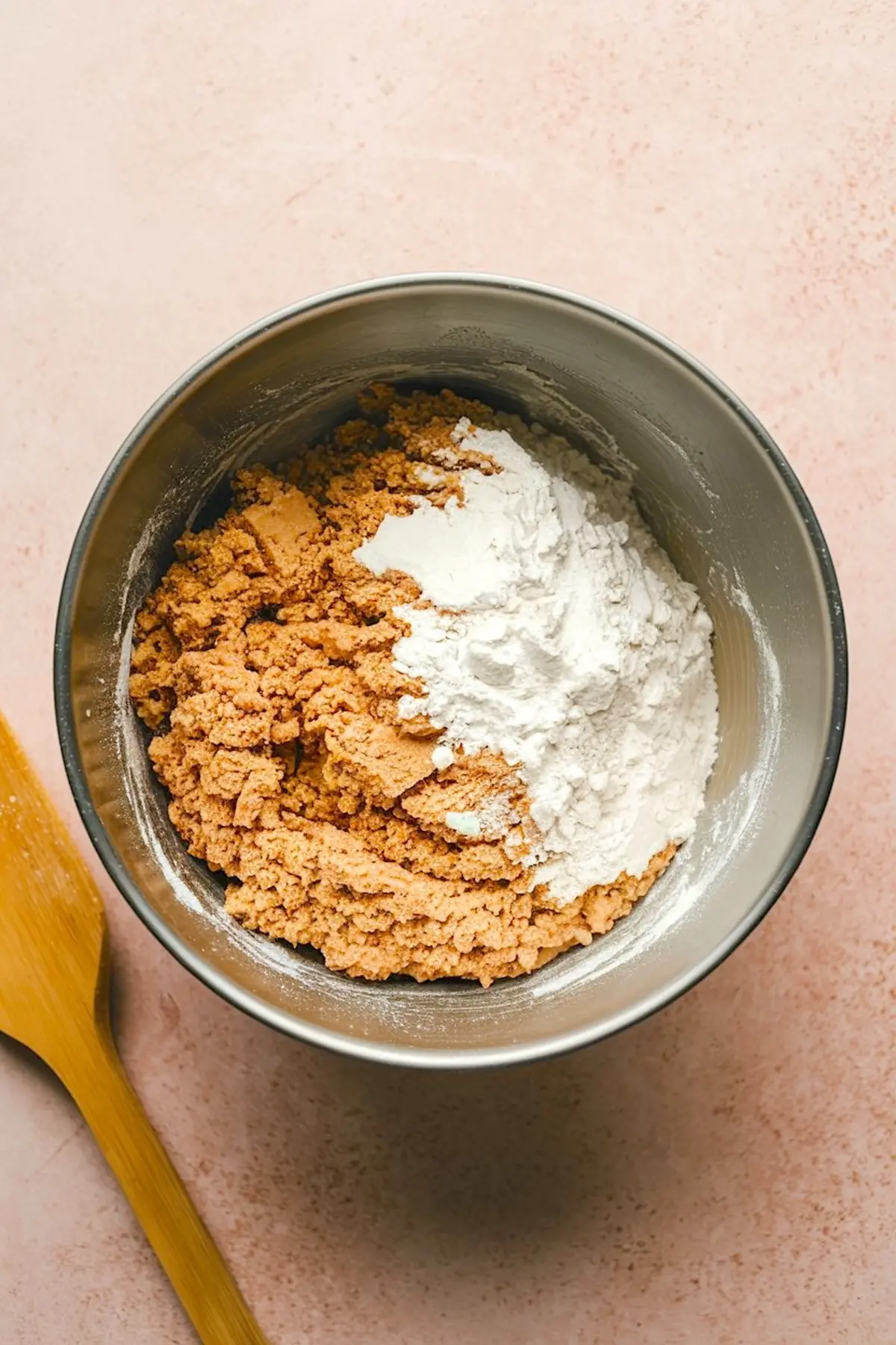 Mixing bowl filled with a partially mixed dough of brown sugar and flour for preparing the base layer of brown butter honey pistachio bars, placed on a pink countertop with a wooden spatula.