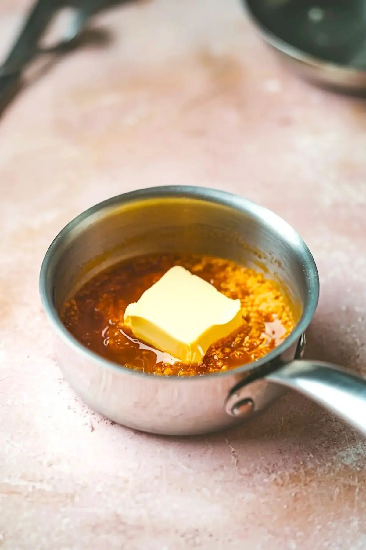Small saucepan containing melting butter and brown sugar mixture for the caramel layer of brown butter honey pistachio bars, set on a pink surface.