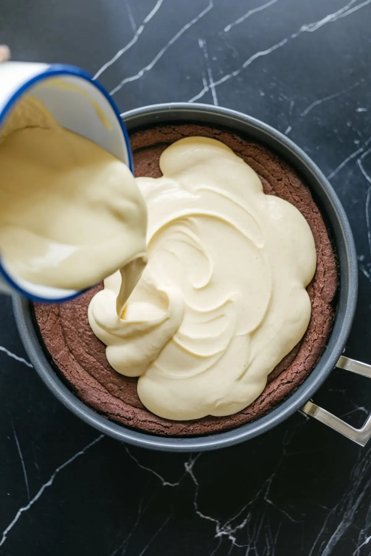 Thick cream cheese filling being poured from a pitcher over a baked brownie crust inside a round springform pan, preparing a layered brownie cheesecake.