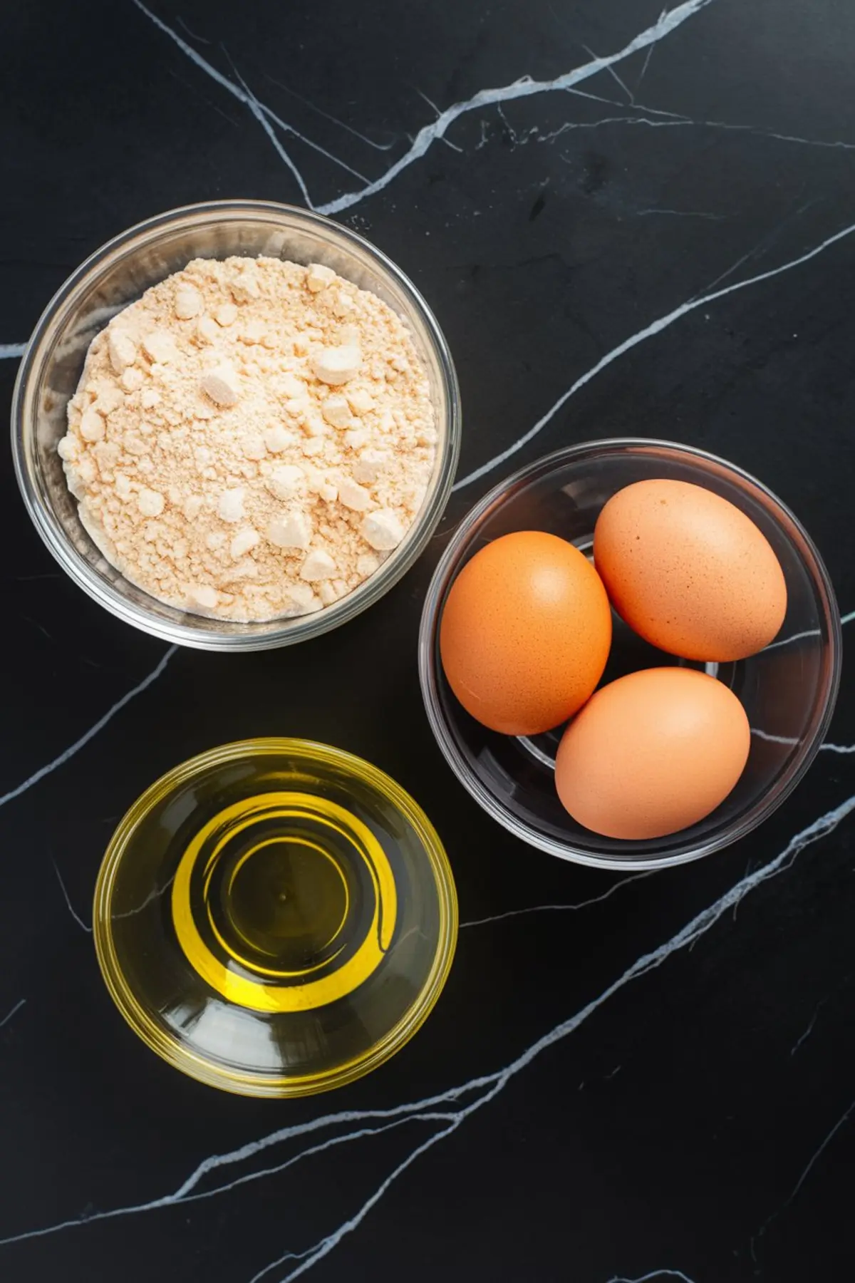 Flat lay showing the three basic ingredients for cake mix cookies: dry cake mix, whole eggs in a bowl, and a small bowl of vegetable oil on a dark marble counter.