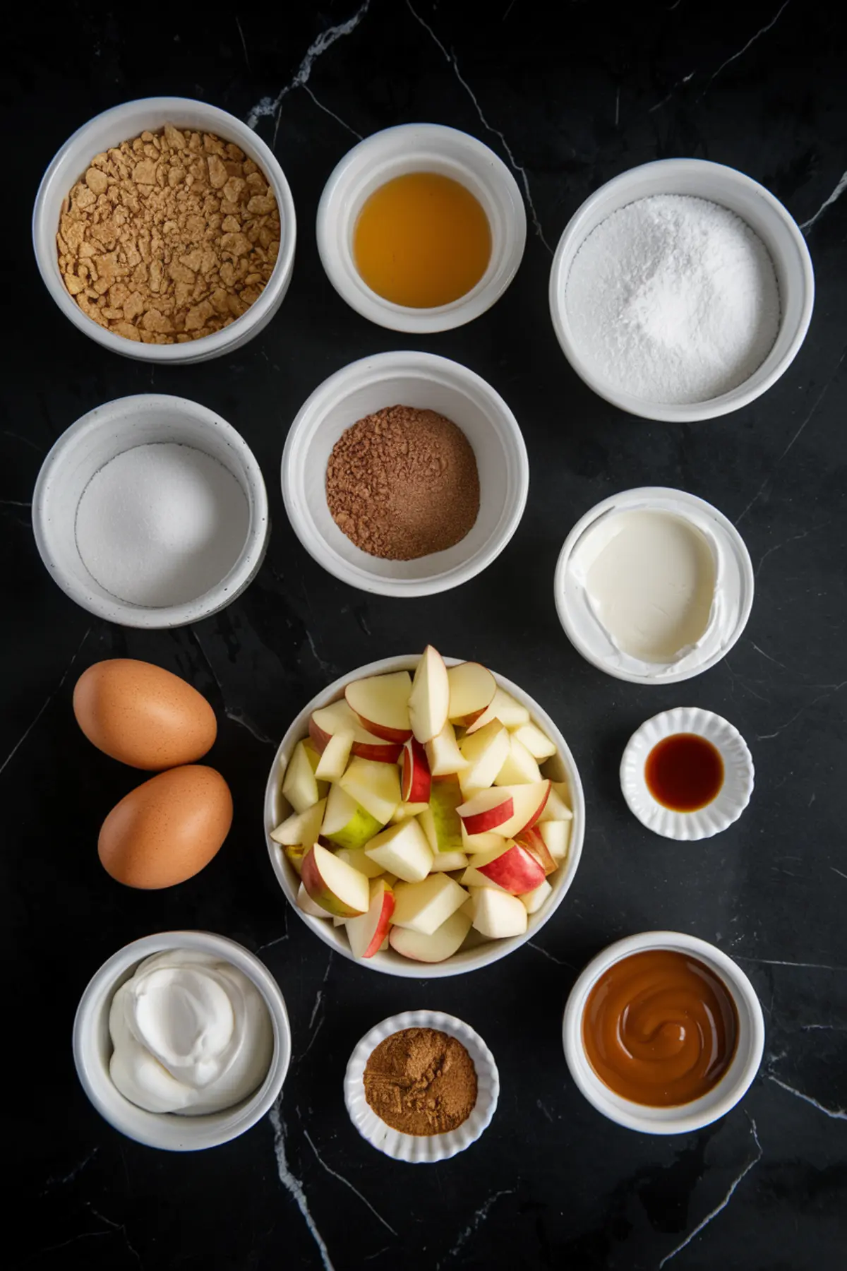 Flat lay of caramel apple cheesecake ingredients including eggs, sugar, cream, vanilla, cinnamon, sour cream, graham crackers, apple chunks, and caramel sauce arranged in white ramekins on a dark surface.