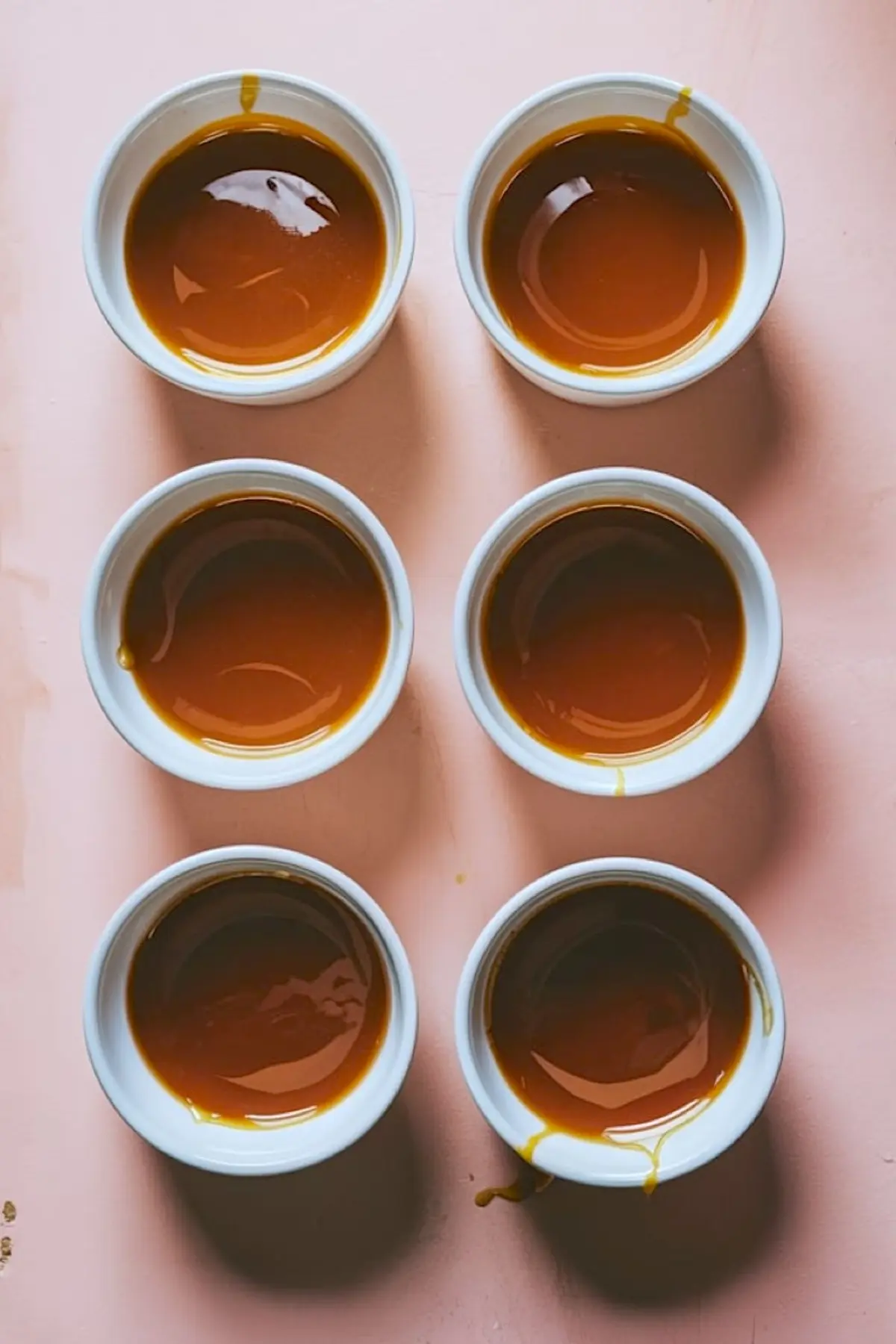 Top view of six white ramekins arranged on a pink surface, each filled with a layer of golden caramel sauce at the bottom.
