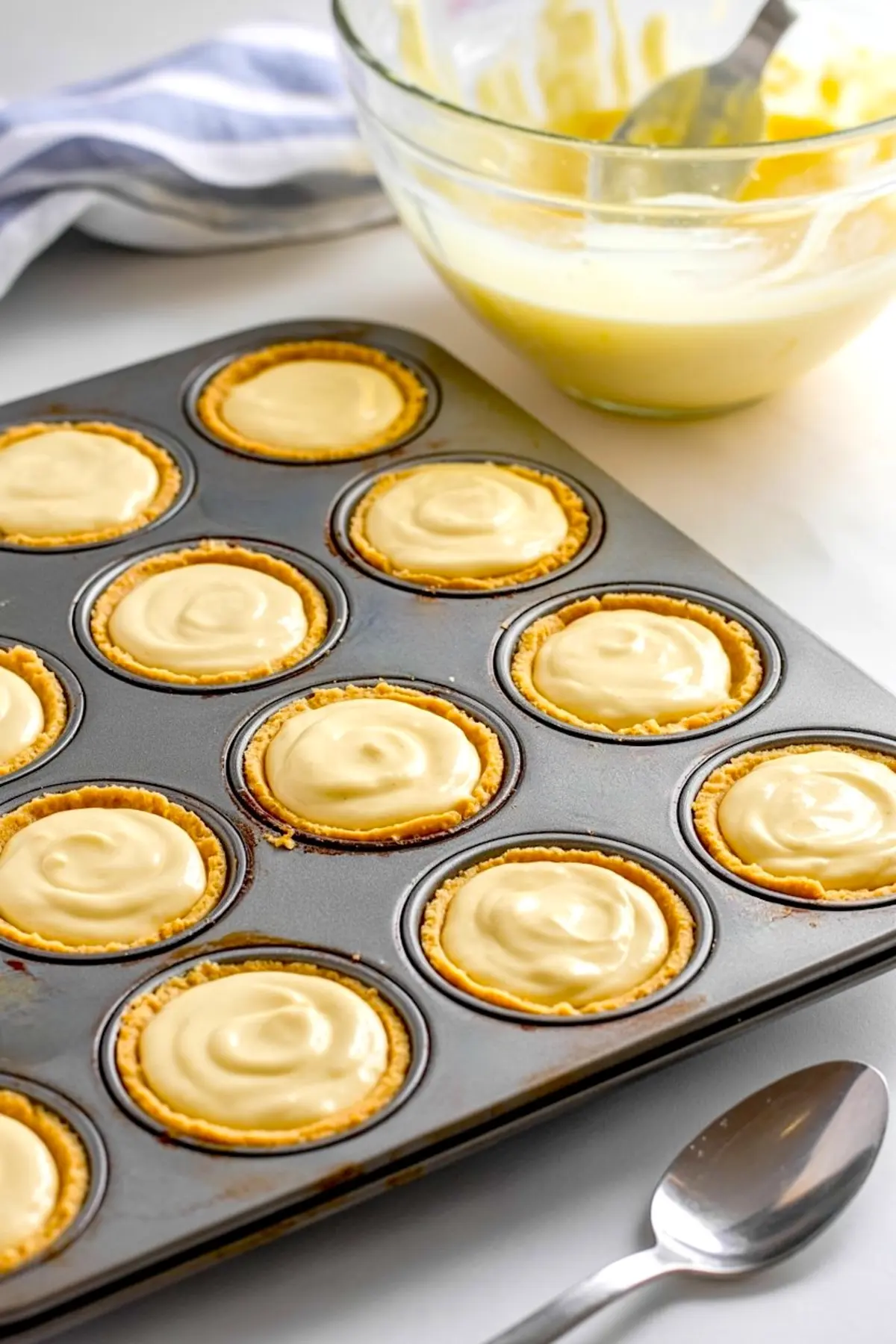 Muffin tin filled with unbaked cheesecake tartlets in golden crusts, with a bowl of batter and spoon in the background on a white counter.