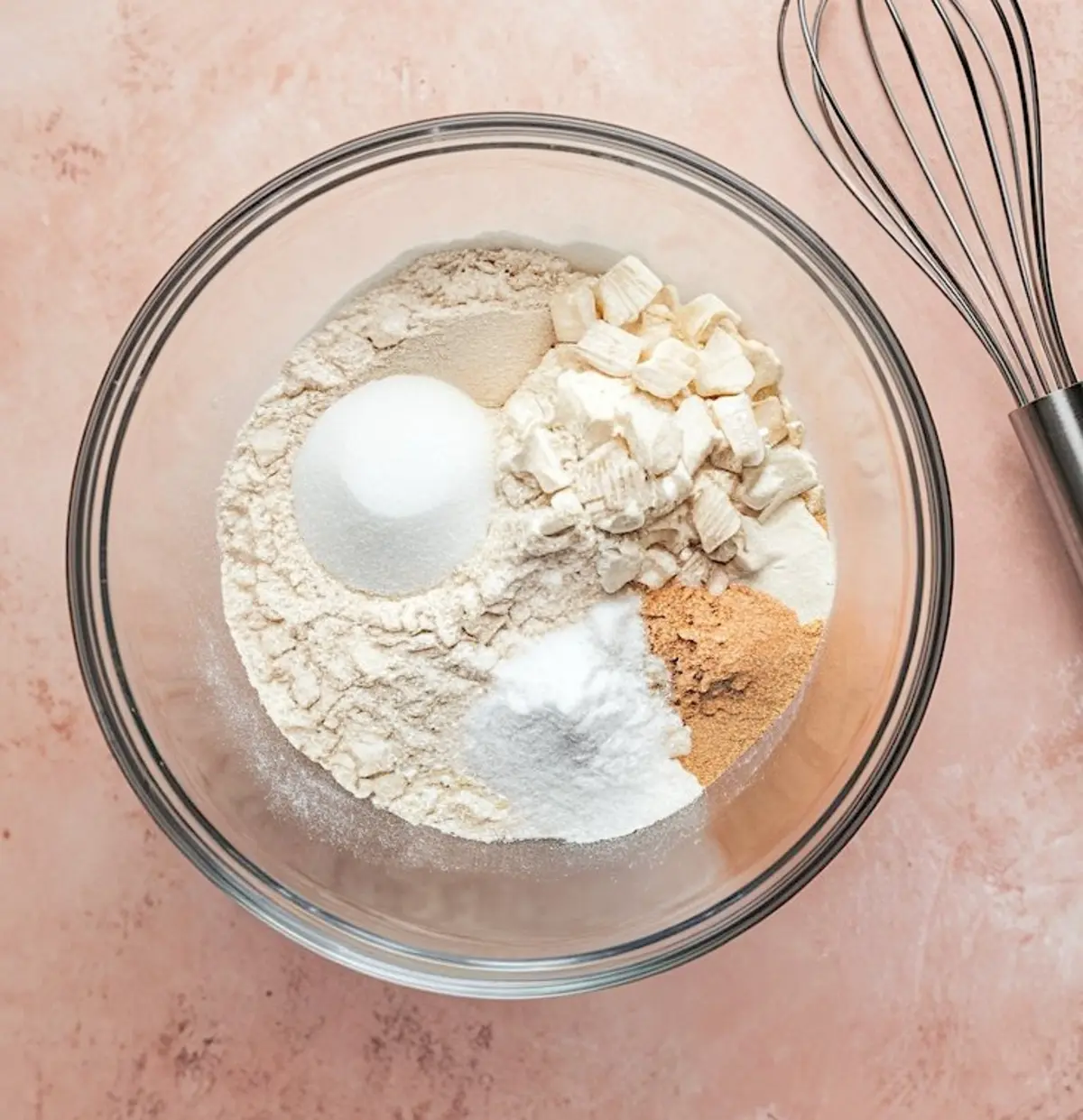 Glass bowl containing measured dry ingredients including flour, sugar, baking powder, and cubed butter, placed next to a wire whisk on a light pink surface.