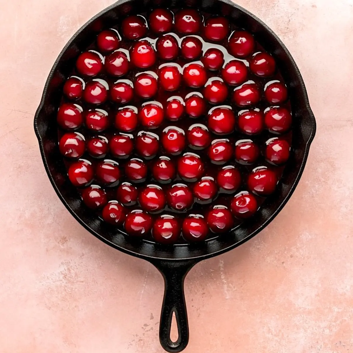 Cast iron skillet filled with glossy, whole red cherries submerged in syrup, prepared as the base layer for homemade cherry cobbler.
