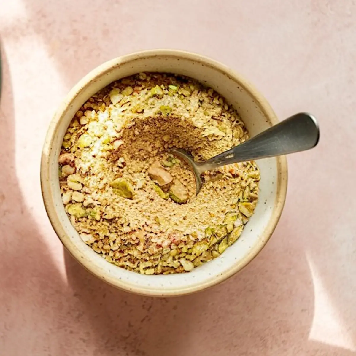 Small ceramic bowl filled with ground pistachio mixture, including visible chopped nuts and fine crumbs, with a metal spoon resting inside, set on a pink background.