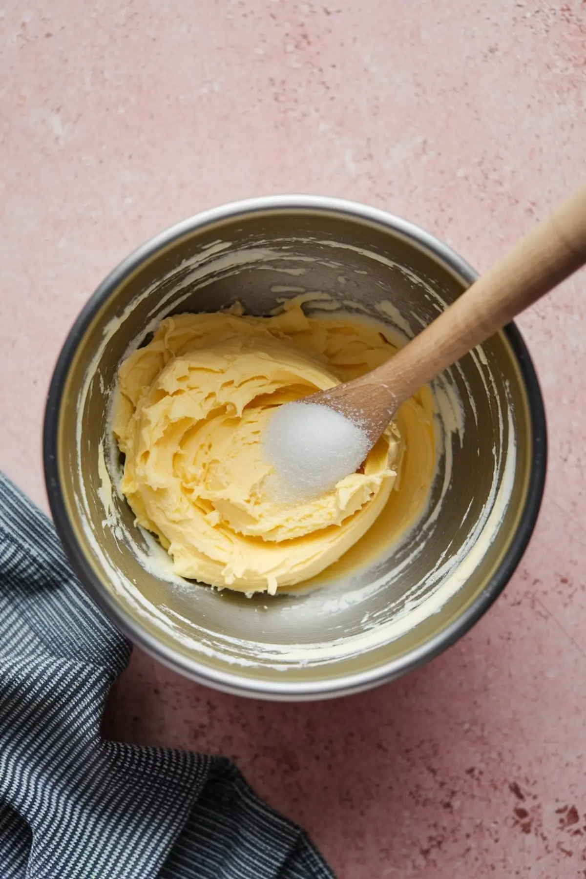 Mixing bowl with whipped butter and sugar, featuring a wooden spoon scooping the creamed mixture, placed on a pink countertop with a striped cloth.