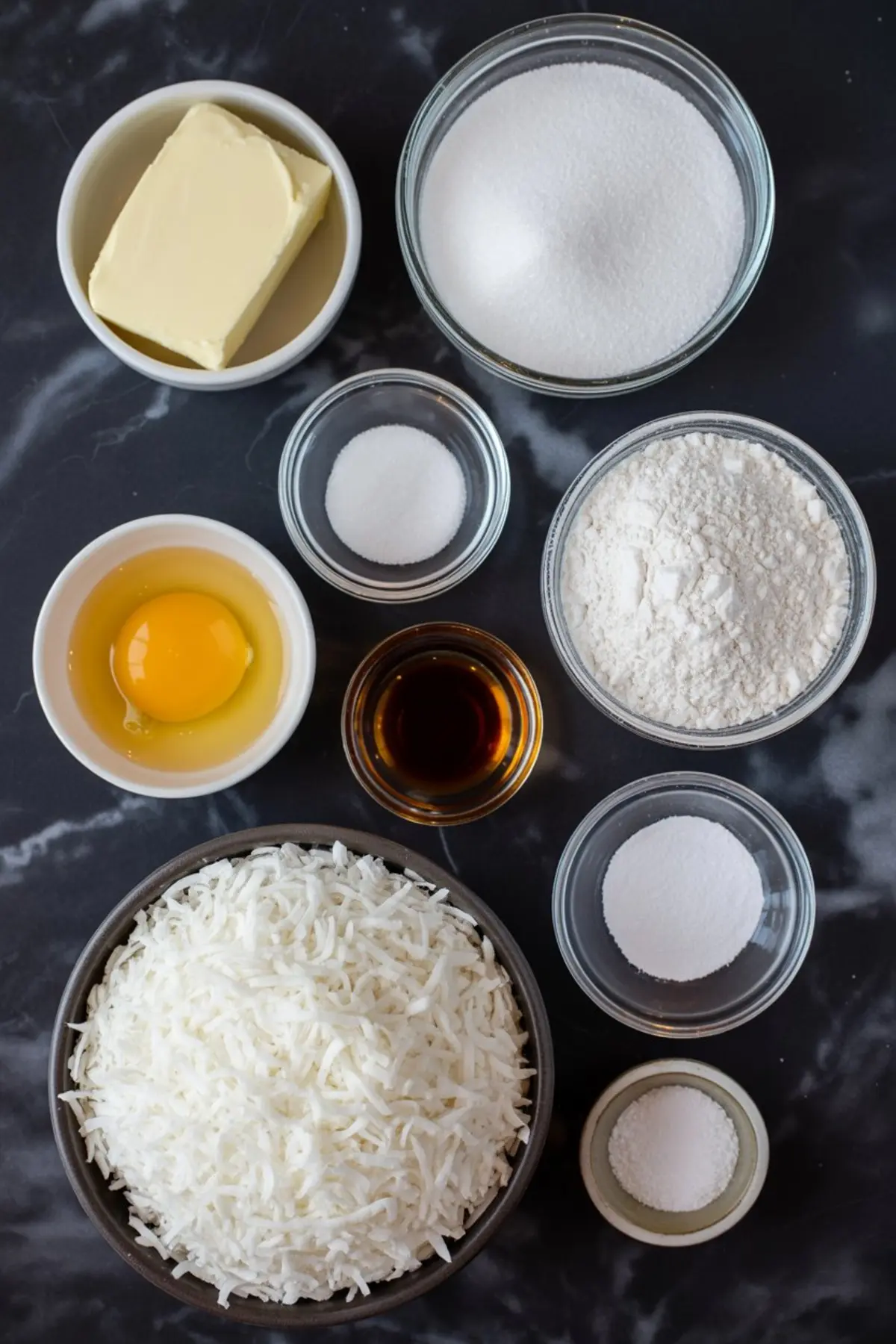Flat lay of coconut cookie ingredients arranged on a dark marble background, including flour, sugar, butter, egg, shredded coconut, vanilla extract, salt, baking soda, and baking powder in small glass and ceramic bowls.