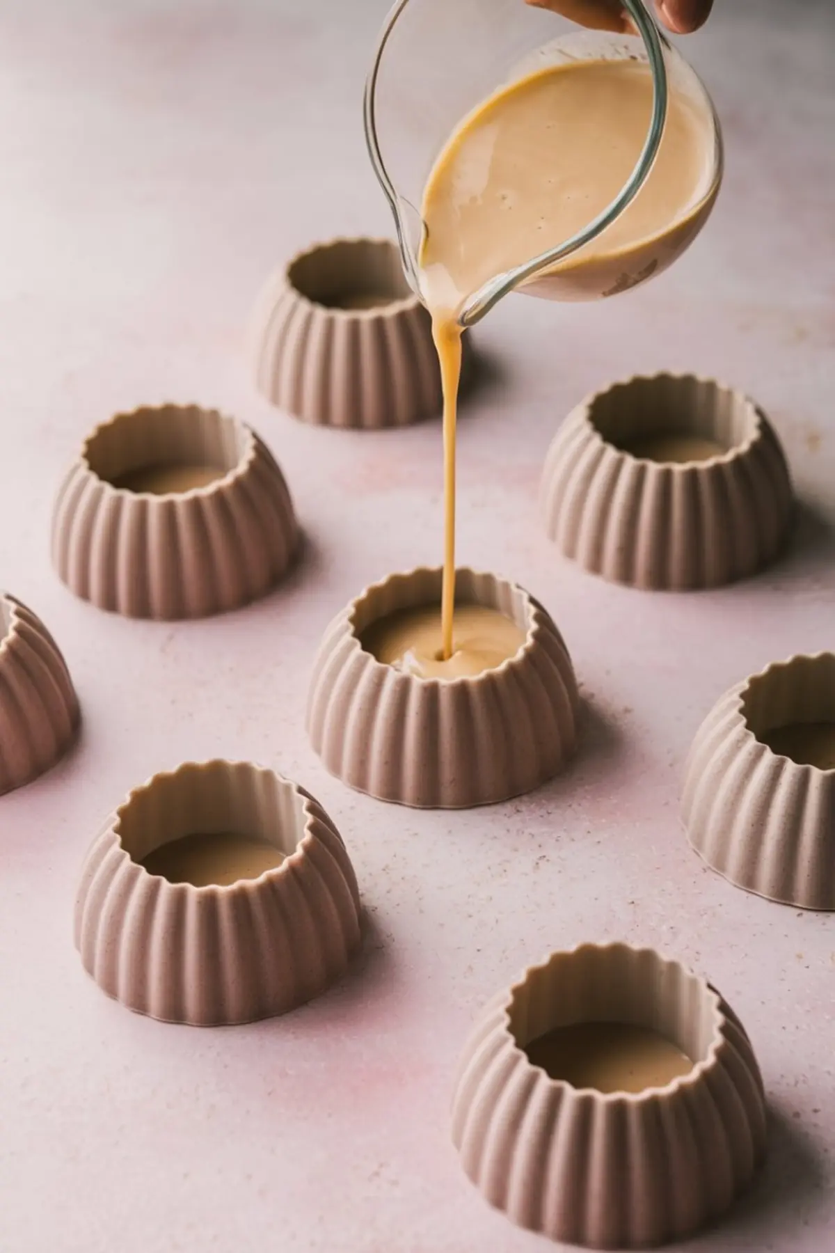 Glass pitcher pouring creamy coffee panna cotta mixture into fluted silicone molds, arranged in rows over a light pink surface for chilling and setting.
