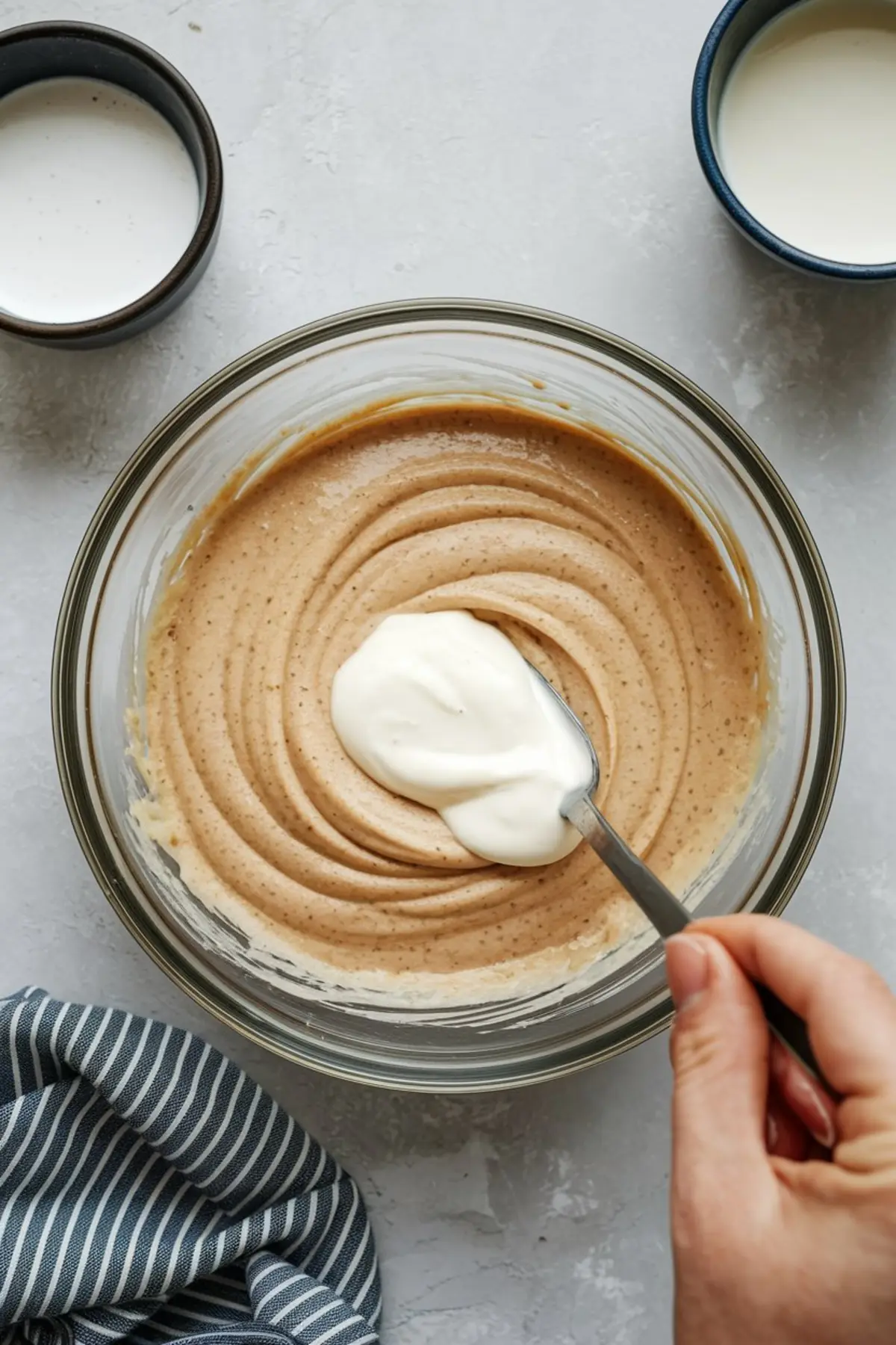 Hand mixing sour cream into light brown cake batter in a glass bowl, surrounded by measuring cups of milk, as part of the preparation for a Labor Day cake or fireworks cake.