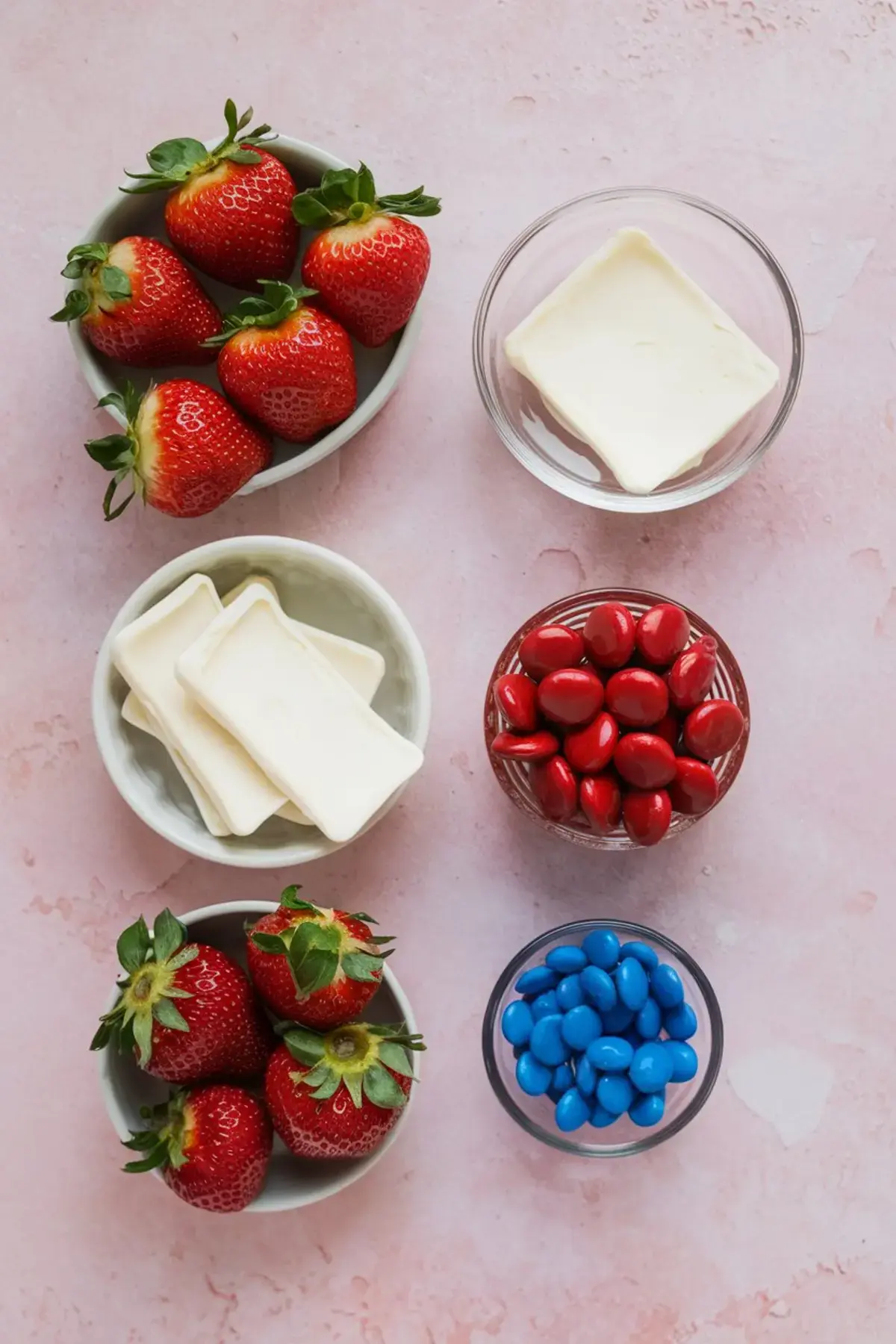 Flat lay of bowls filled with fresh strawberries, white candy melts, white chocolate squares, red candy-coated chocolates, and blue candy-coated chocolates on a pink background.
