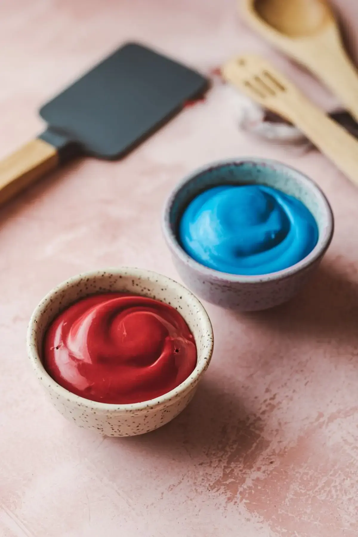 Two small ceramic bowls filled with red and blue colored melted candy coating set on a pink surface with wooden kitchen utensils in the background.
