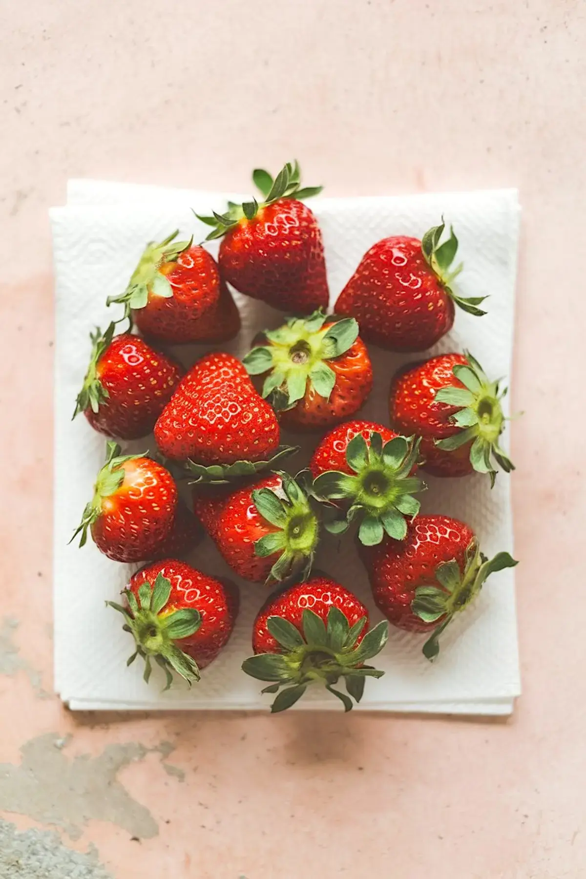 Group of fresh whole strawberries with green stems arranged on a white paper towel over a pink textured surface.
