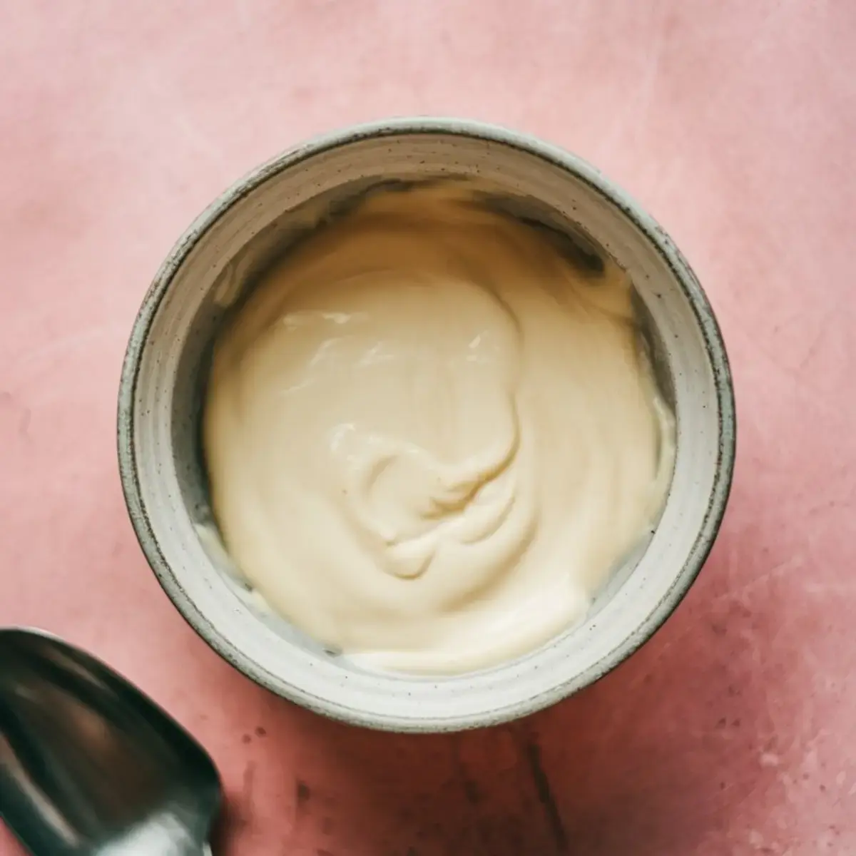 Bowl of smooth, melted white chocolate with a metal spoon beside it on a pink surface.
