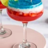 Close-up of a tricolor frozen margarita with red strawberry base, white coconut layer, and blue top, decorated with a lime slice and miniature U.S. flag in a festive cocktail glass.