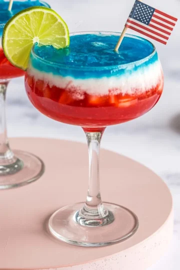 Close-up of a tricolor frozen margarita with red strawberry base, white coconut layer, and blue top, decorated with a lime slice and miniature U.S. flag in a festive cocktail glass.