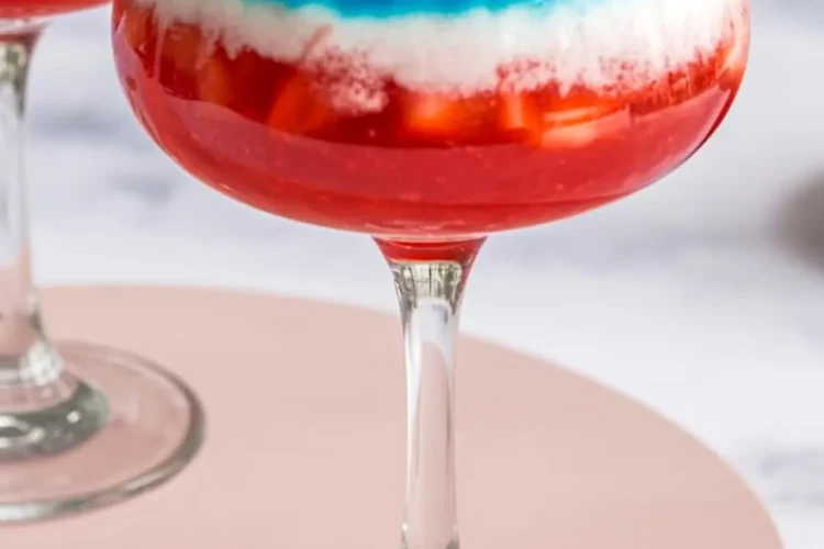 Close-up of a tricolor frozen margarita with red strawberry base, white coconut layer, and blue top, decorated with a lime slice and miniature U.S. flag in a festive cocktail glass.