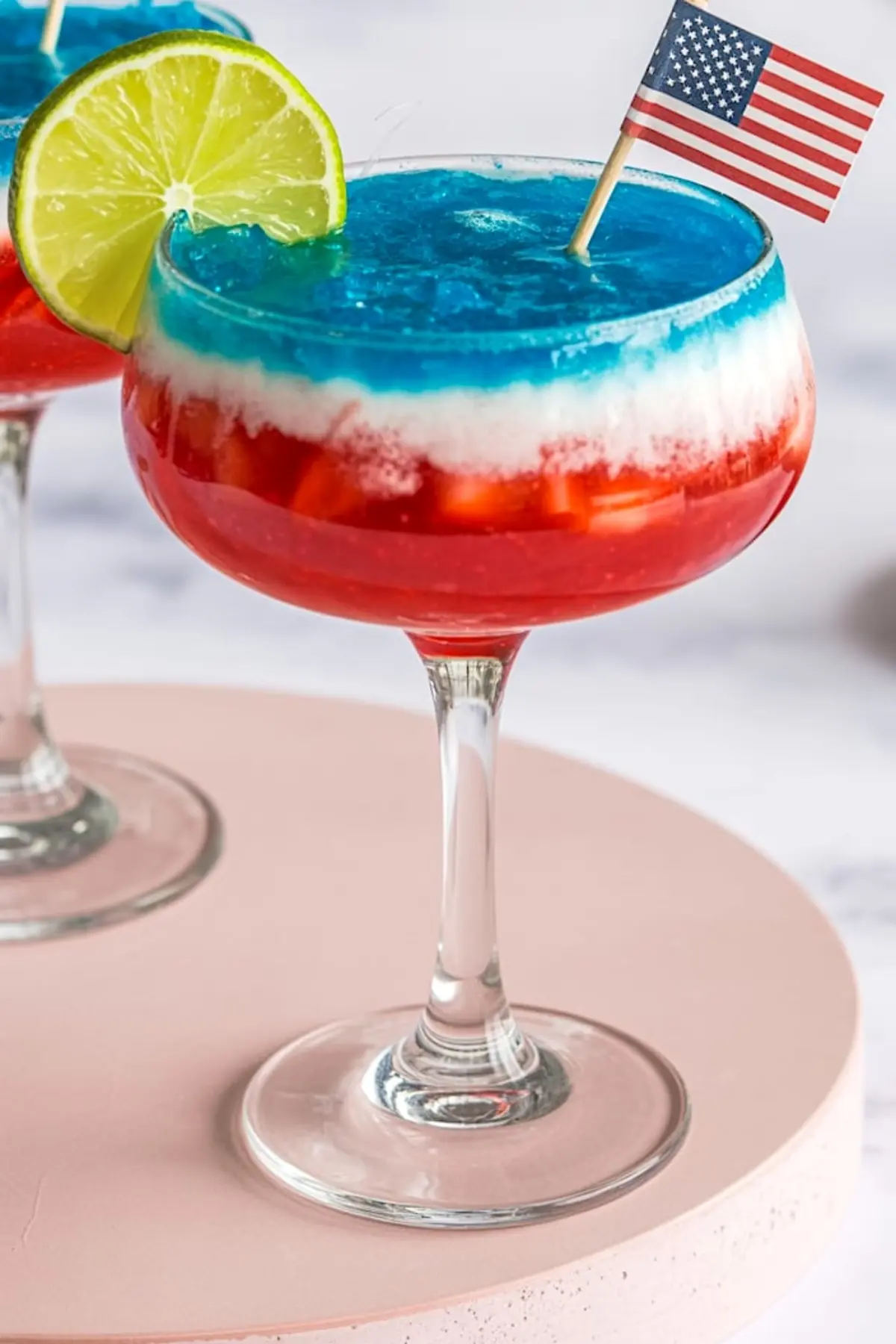 Close-up of a tricolor frozen margarita with red strawberry base, white coconut layer, and blue top, decorated with a lime slice and miniature U.S. flag in a festive cocktail glass.
