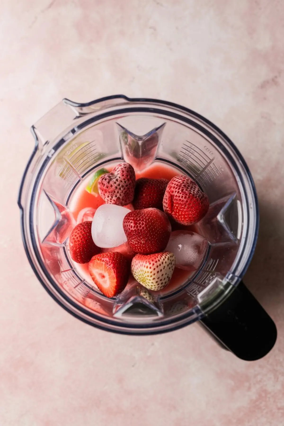 Overhead view of a blender filled with strawberries, ice cubes, and red liquid mixture, preparing the red layer for a layered frozen margarita recipe.
