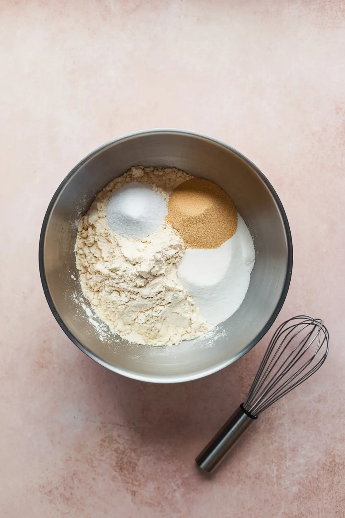 Mixing bowl filled with dry ingredients for butter mochi, including rice flour, sugar, baking powder, and salt, next to a stainless steel whisk on a pink background.