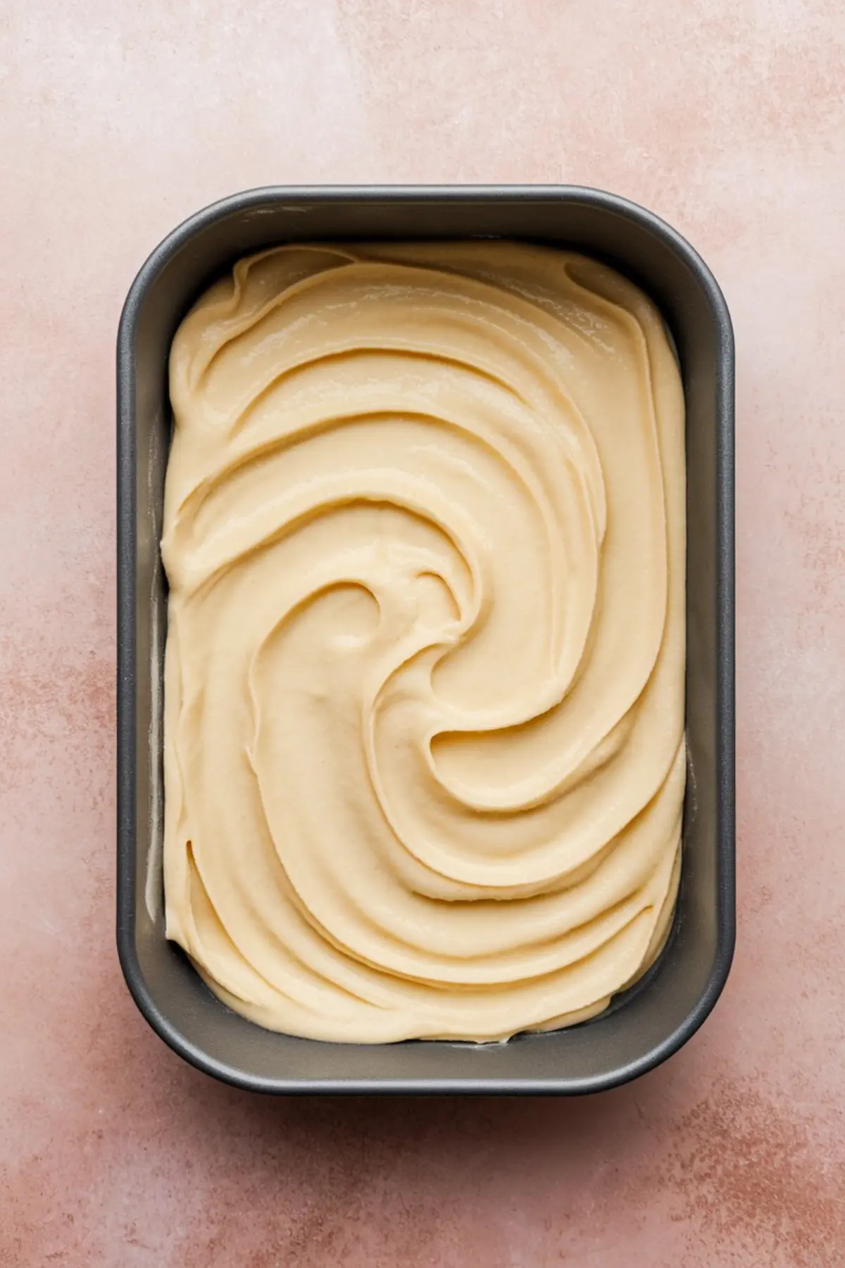 Overhead view of mochi batter swirled in a metal baking pan, showing thick, creamy, golden batter texture before baking for traditional Hawaiian butter mochi.