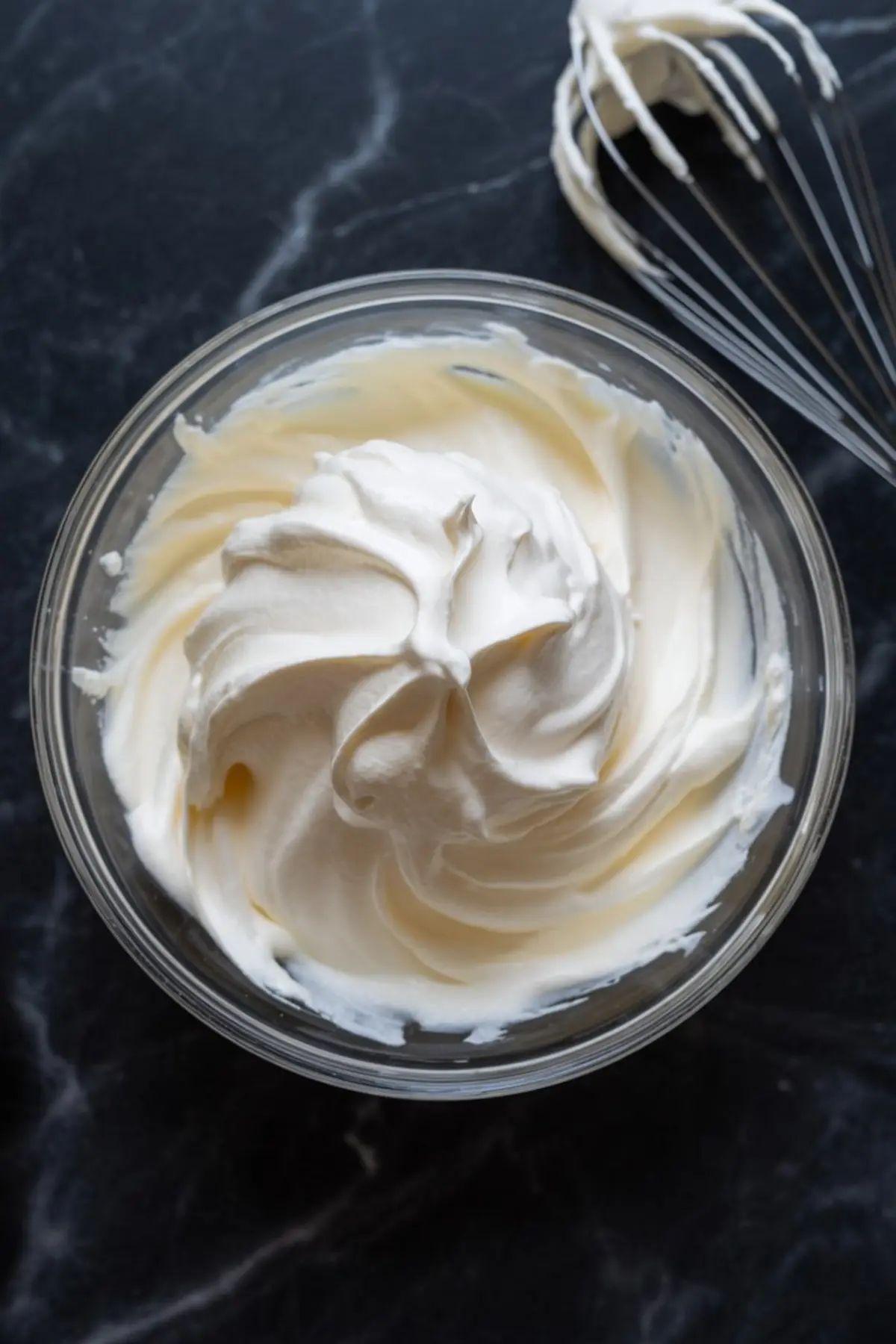 Thick whipped cream swirled in a glass bowl with a metal whisk on a dark marble countertop, highlighting a soft, airy texture for homemade ice cream base.